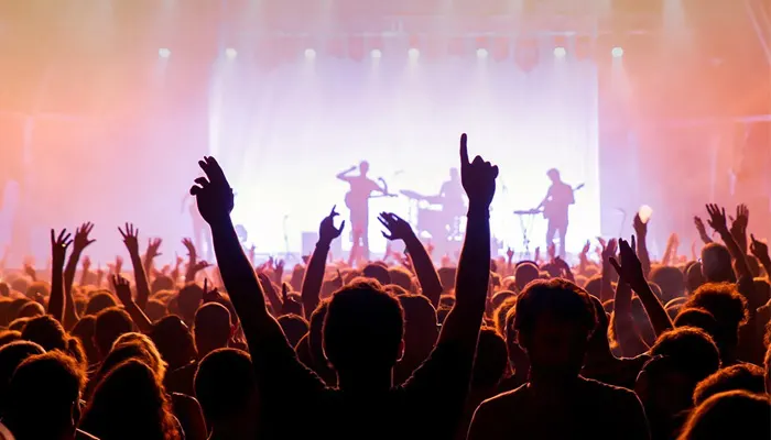 Folla a un concerto con mani alzate. Il palco illuminato mostra sagome di musicisti con strumenti.