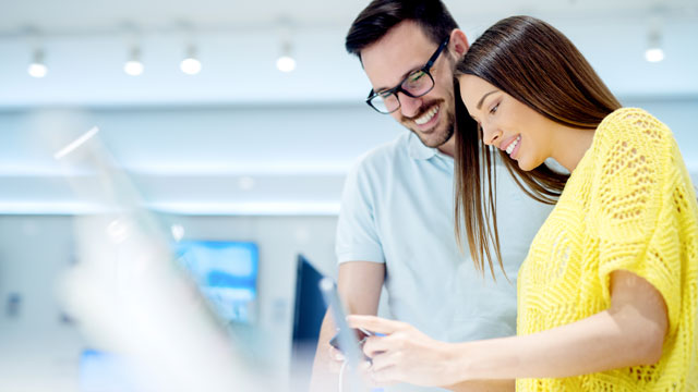 Donna e uomo guardano un telefono in un negozio. Lei indossa una camicetta gialla; lui una polo azzurra e occhiali.