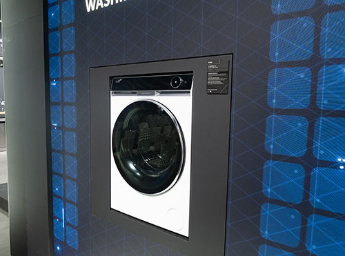 Lavatrice bianca e nera incassata in una parete grigia con motivo a riquadri blu. Pannello di controllo visibile.