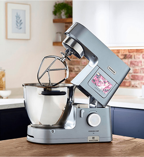 Macchina da cucina grigia con ciotola in acciaio e frusta, su un tavolo di legno in una cucina con mattoni a vista.