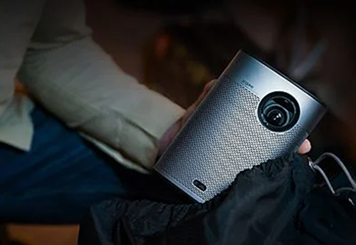 Un proiettore portatile argento è tenuto in mano, con un obiettivo nero visibile. Lo sfondo è sfocato, con tessuti chiari e scuri.