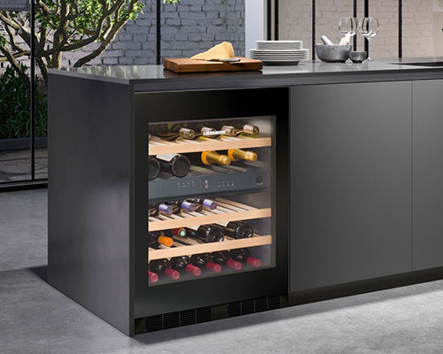 Cucina grigia con cantinetta vini a ripiani in legno, bottiglie di vino e formaggio su tagliere. Piante e vetrate sullo sfondo.