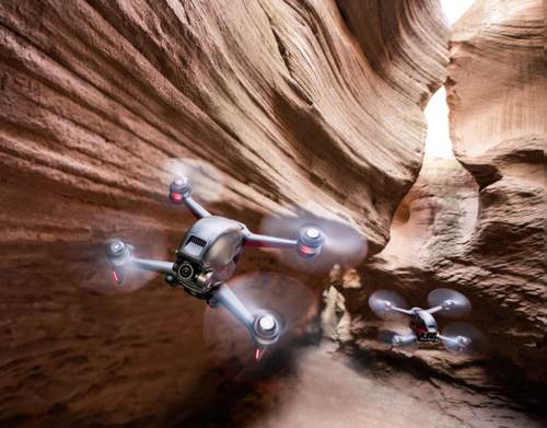 Due droni grigi volano in un canyon di roccia. Le pareti del canyon sono alte e strette, con strati orizzontali.