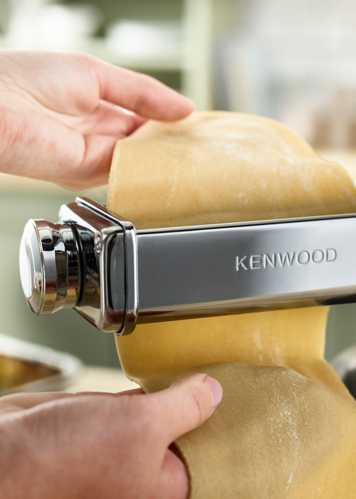Una sfoglia di pasta passa attraverso una macchina per la pasta Kenwood. Le mani guidano la pasta attraverso i rulli metallici.