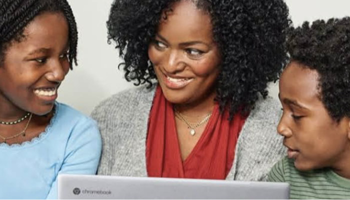 Madre e due figli guardano un laptop. La madre indossa una sciarpa rossa e una giacca grigia. I figli indossano abiti blu e verdi.