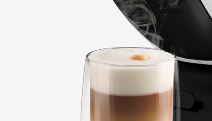Bicchiere di bevanda al latte con strati di caffè e schiuma, accanto a una macchina da caffè nera.