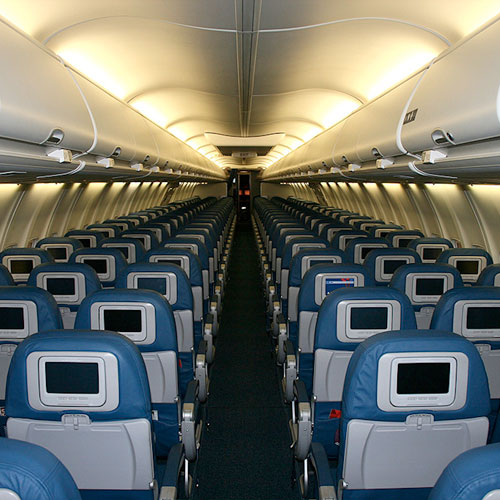 Interno di un aereo con sedili blu disposti in file su moquette scura e schermi grigi sui poggiatesta.