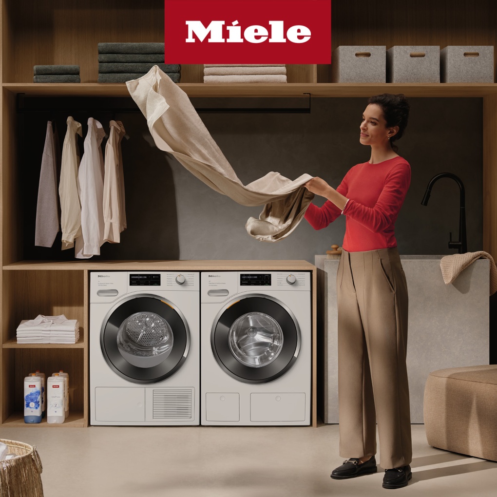 una donna che controlla dei vestiti e sullo sfondo due lavatrici e scaffali conteneti altri panni. In alto logo Miele
