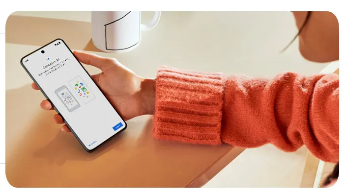 Una mano tiene un telefono con una schermata bianca. Sullo sfondo ci sono una tazza bianca e una superficie marrone chiaro.