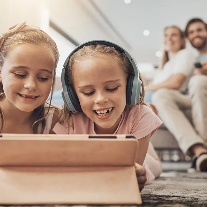 Due bambine usano un tablet, una con cuffie blu, con due adulti sfocati sullo sfondo.