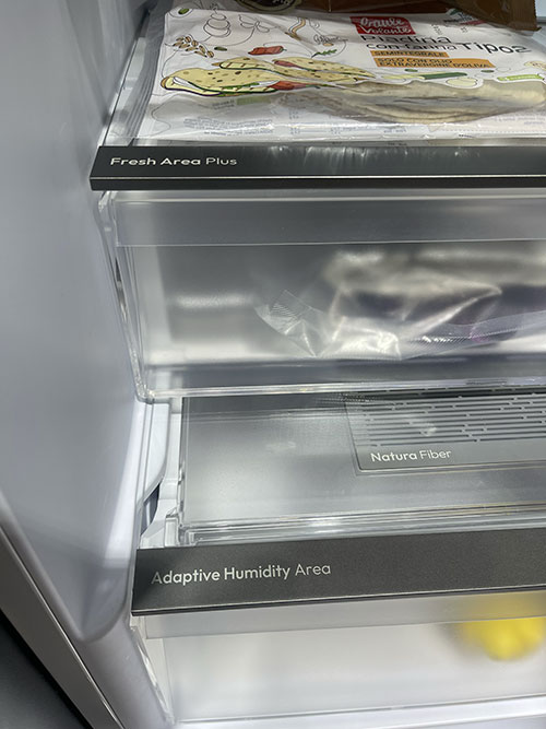 Ripiano del frigorifero con confezione di cibo sopra. Cassetti trasparenti etichettati: "Fresh Area Plus", "Natura Fiber", "Adaptive Humidity Area".