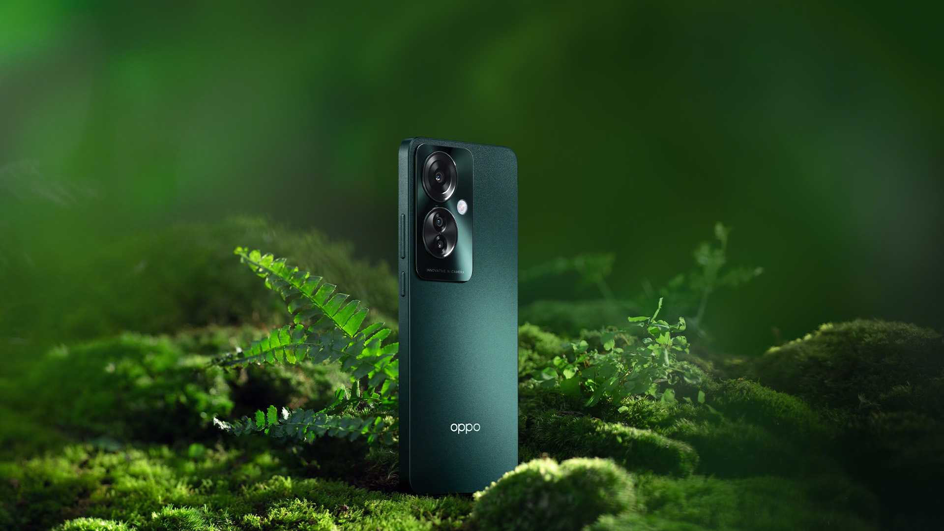 Un telefono verde scuro con due fotocamere poggia su muschio verde brillante con una felce e vegetazione sullo sfondo.