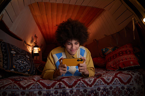 Ragazzo su letto con cuscini decorati, usa un telefono giallo. Lampada e luci decorative sullo sfondo.