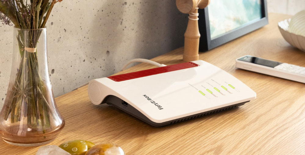 Su un tavolo di legno chiaro, un router bianco e rosso, un telefono bianco, un vaso con fiori secchi e altri oggetti.