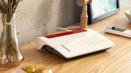 Su un tavolo di legno chiaro, un router bianco e rosso, un telefono bianco, un vaso con fiori secchi e altri oggetti.