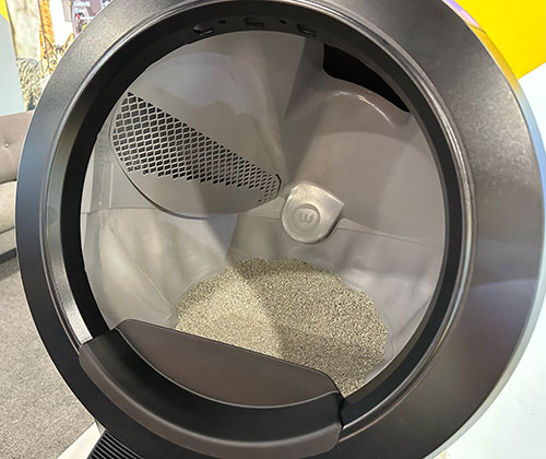 Vista ravvicinata di una lettiera automatica rotonda con sassolini grigi all'interno e un ingresso scuro.