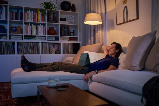 Uomo disteso su un divano bianco con un laptop. Una libreria piena e una lampada sono sullo sfondo.