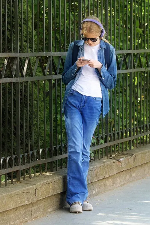 Donna con cuffie e occhiali da sole usa un telefono. Indossa jeans, camicia bianca e giacca di jeans. Dietro, recinzione metallica e alberi.