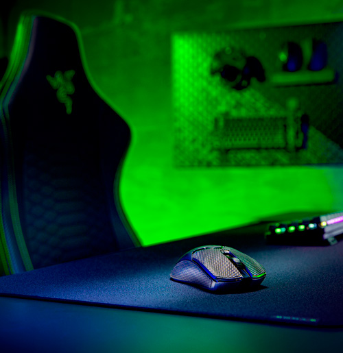 Mouse grigio su tappetino blu, con sedia gaming nera e luci verdi sullo sfondo. Tastiera visibile parzialmente.