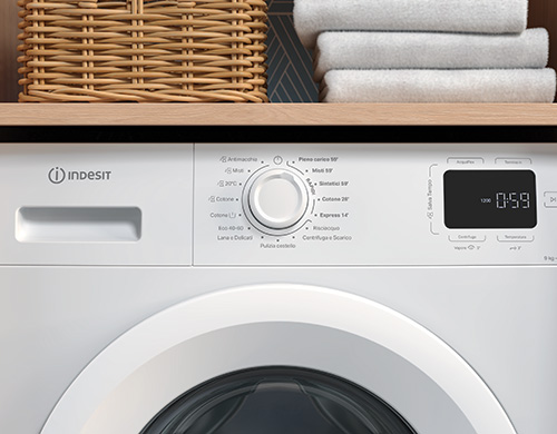 Primo piano di una lavatrice bianca Indesit, con cesto di vimini e asciugamani bianchi impilati sullo scaffale. Sul display si legge "0:59".