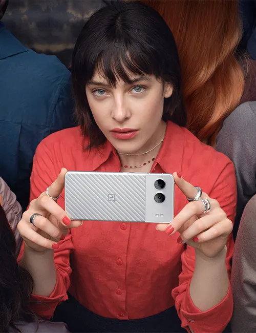 Donna in camicia rossa tiene un telefono orizzontale. Ha anelli alle dita. Sullo sfondo, altre persone.