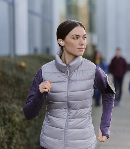 Donna con auricolari che corre all'aperto. Indossa un gilet imbottito grigio sopra una maglia viola a maniche lunghe.