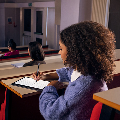 Aula con studenti. Una studentessa scrive su un tablet con una penna. Banchi in legno, sedili rossi. Sullo sfondo, altri studenti.