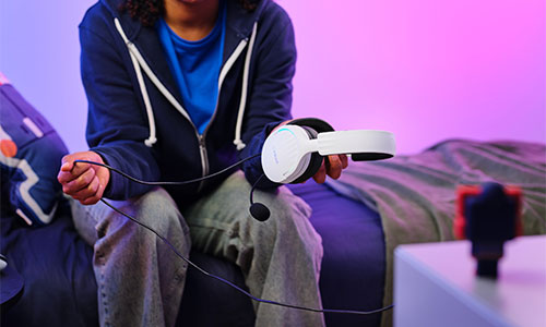 Persona seduta su un letto tiene cuffie bianche. Sul comodino un supporto per telefono. Sfondo viola e blu.