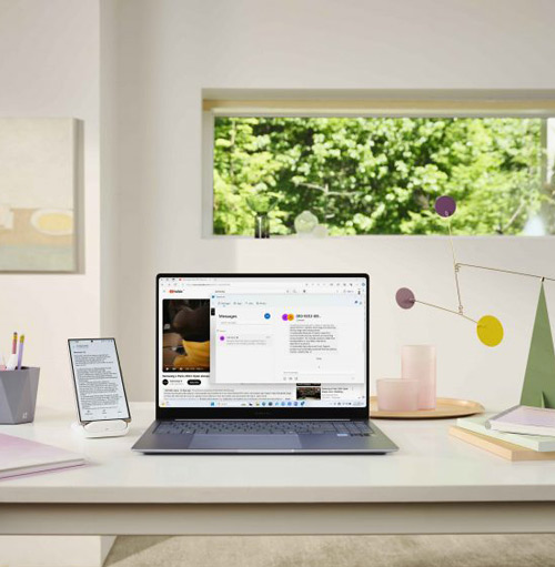 Scrivania bianca con laptop aperto, tablet, oggetti decorativi. Finestra con vista sul verde. Illuminazione chiara e naturale.