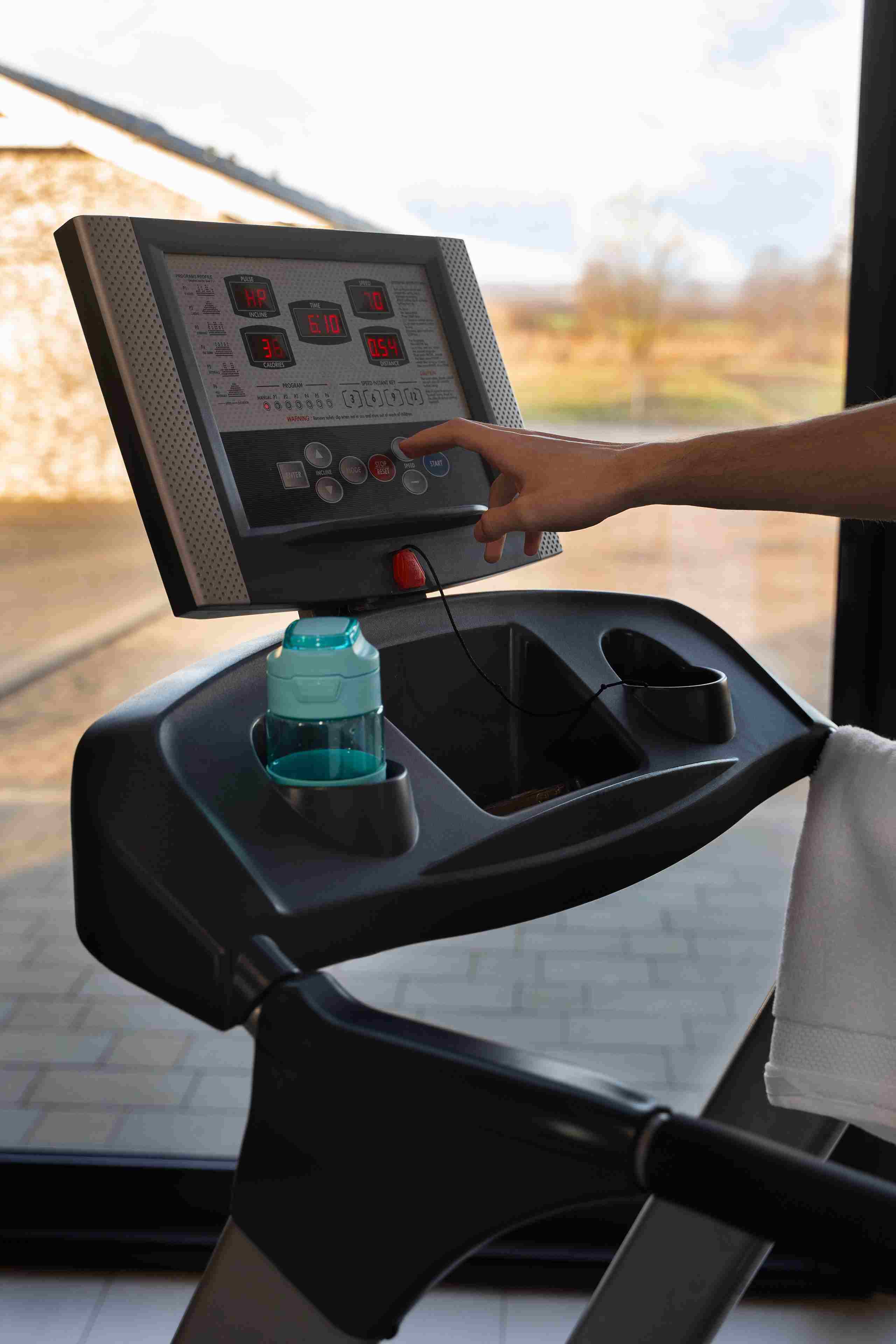 Tapis roulant nero con schermo che mostra dati di allenamento. Una mano preme il pulsante start. Bottiglia d'acqua e asciugamano visibili.