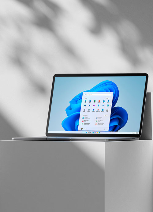 Un laptop grigio aperto mostra il sistema operativo Windows su uno sfondo blu chiaro, posizionato su un cubo bianco.