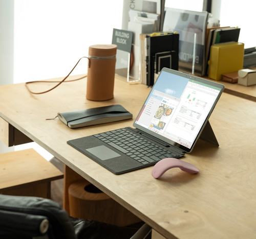 Scrivania con laptop aperto, mouse rosa, custodia grigia, speaker cilindrico marrone chiaro e libri sullo sfondo.