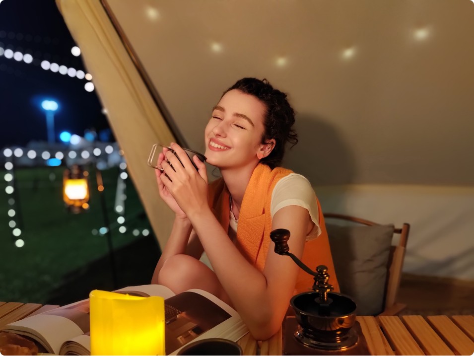 Donna sorridente beve da tazza in tenda con libro, candela gialla e macinino da caff� su tavolo di legno. All'esterno luci notturne.