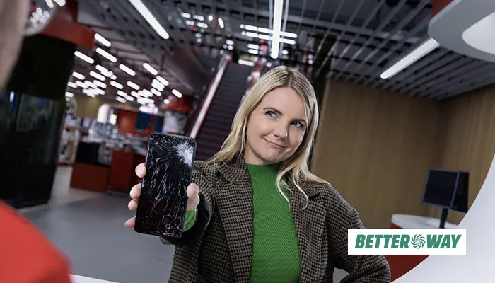 Donna in negozio mostra cellulare rotto. Scala mobile e luci a soffitto sullo sfondo.