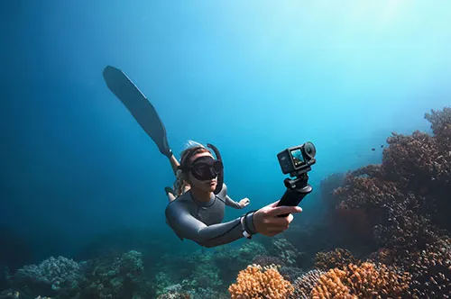 Una persona in tuta da sub con pinne e maschera nuota vicino al corallo, tenendo una action cam. Acqua blu sullo sfondo.