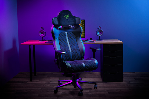 Sedia gaming nera e blu con logo verde su sfondo viola e blu. Scrivania di legno con altoparlanti e armadietto nero.