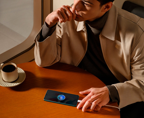 Uomo seduto a un tavolo di legno con tazza di caff� e smartphone in carica, vicino a una finestra.