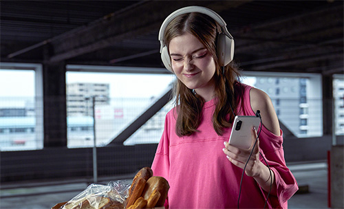 Donna con cuffie bianche, maglia rosa, tiene cellulare e pane. Sfondo urbano con finestre.
