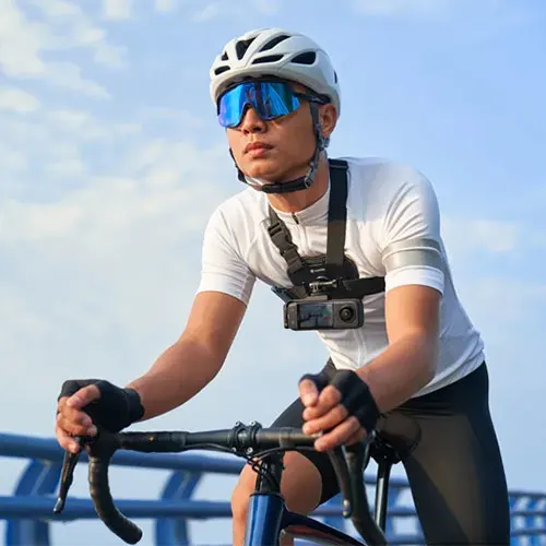 Ciclista con casco bianco, occhiali blu e maglia bianca. Indossa una action camera sul petto. In sella a una bici blu su un ponte.
