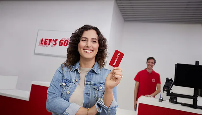 Donna sorridente con giacca di jeans che mostra una carta rossa. Uomo in maglietta rossa in piedi dietro un bancone.