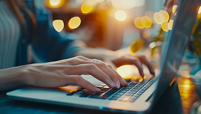 Mani che digitano su una tastiera di laptop grigia. Sfondo sfocato con luci gialle.
