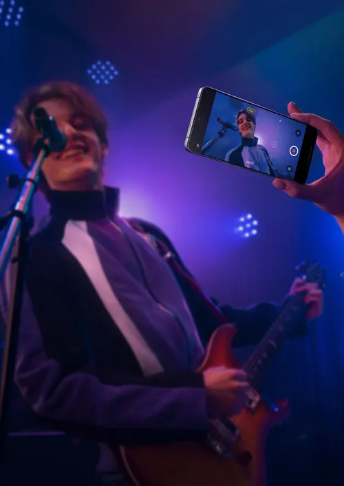 Un uomo suona la chitarra e canta al microfono su un palco illuminato. Un telefono riprende l'uomo che canta.