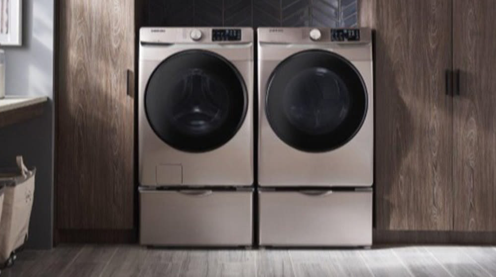 Due lavatrici grigie affiancate con obl� scuro e cassetti inferiori, su pavimento grigio. Armadi in legno sullo sfondo.