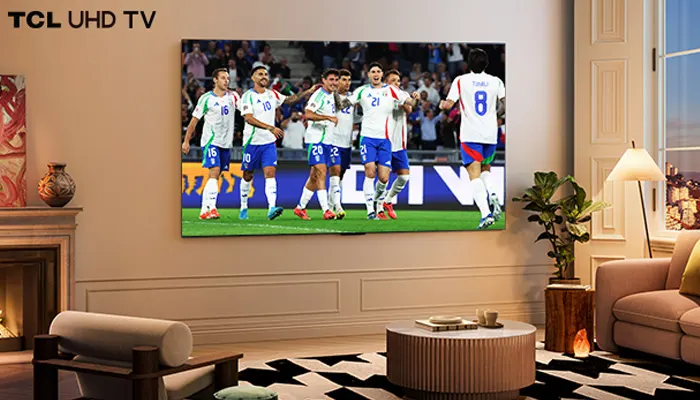 Soggiorno con TV che mostra una partita di calcio. Divano beige, lampada da terra, camino e tappeto geometrico in bianco e nero.