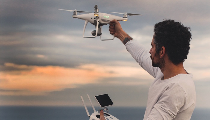 Un uomo di profilo tiene un drone bianco sollevato in aria con una mano e il radiocomando con l'altra, sullo sfondo di un cielo nuvoloso al tramonto sopra il mare.