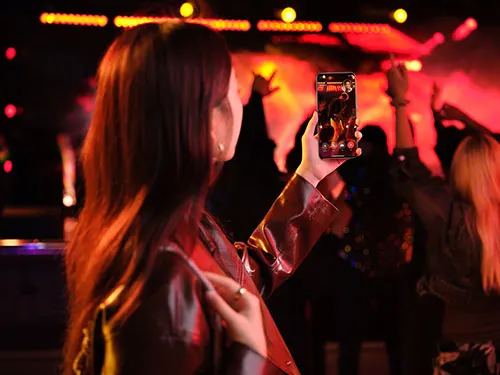 Donna con cellulare in concerto illuminato di rosso. Persone ballano sullo sfondo.