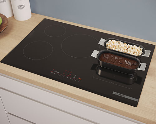 Piano cottura nero con pentole: una contiene popcorn e l'altra una torta al cioccolato. Sul ripiano ci sono barattoli bianchi.