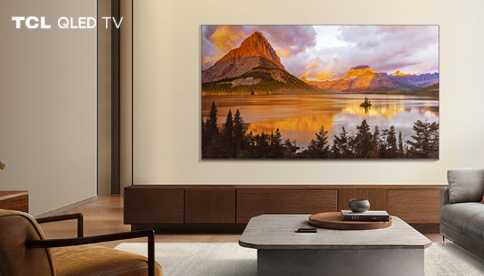 Un televisore TCL QLED TV installato a parete sopra un lungo mobile in legno scuro in un salotto moderno. Sullo schermo è visualizzato un paesaggio montano con un lago che riflette le luci calde del tramonto.