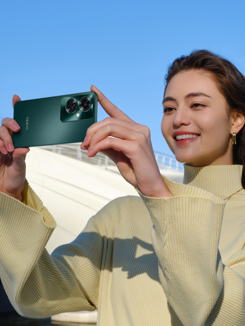Donna sorridente fotografa con smartphone verde. Indossa maglia beige a maniche lunghe. Cielo azzurro e struttura bianca sullo sfondo.