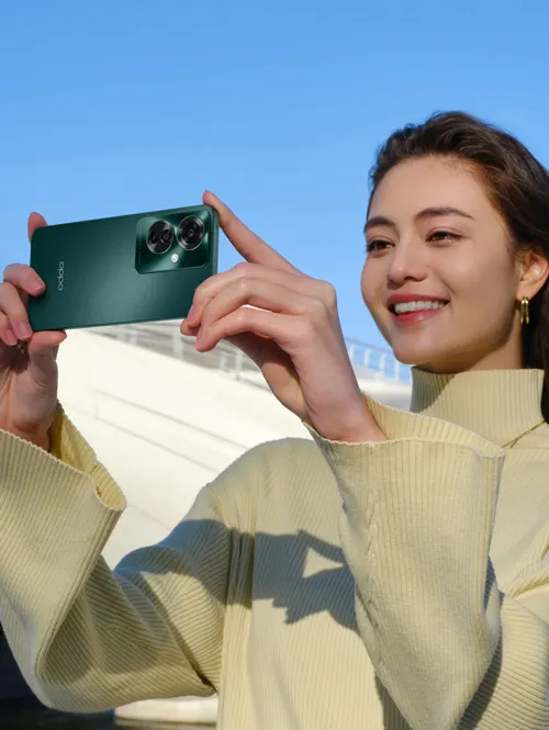 Donna sorridente fotografa con smartphone verde. Indossa maglia beige a maniche lunghe. Cielo azzurro e struttura bianca sullo sfondo.